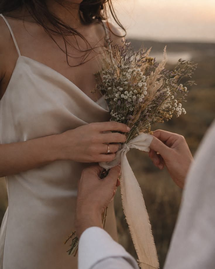 A bride in an ivory slip dress holding a bouquet of dried wildflowers as her partner ties the ribbon