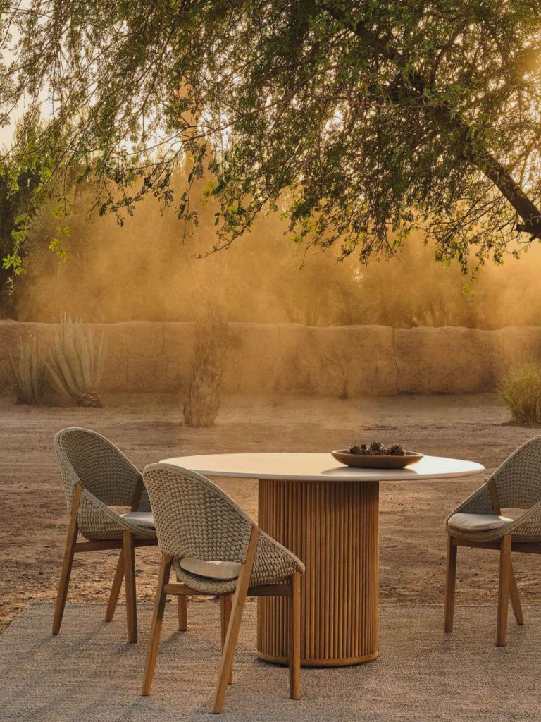 A round fluted timber table and woven chairs set beneath a tree in a sunlit garden