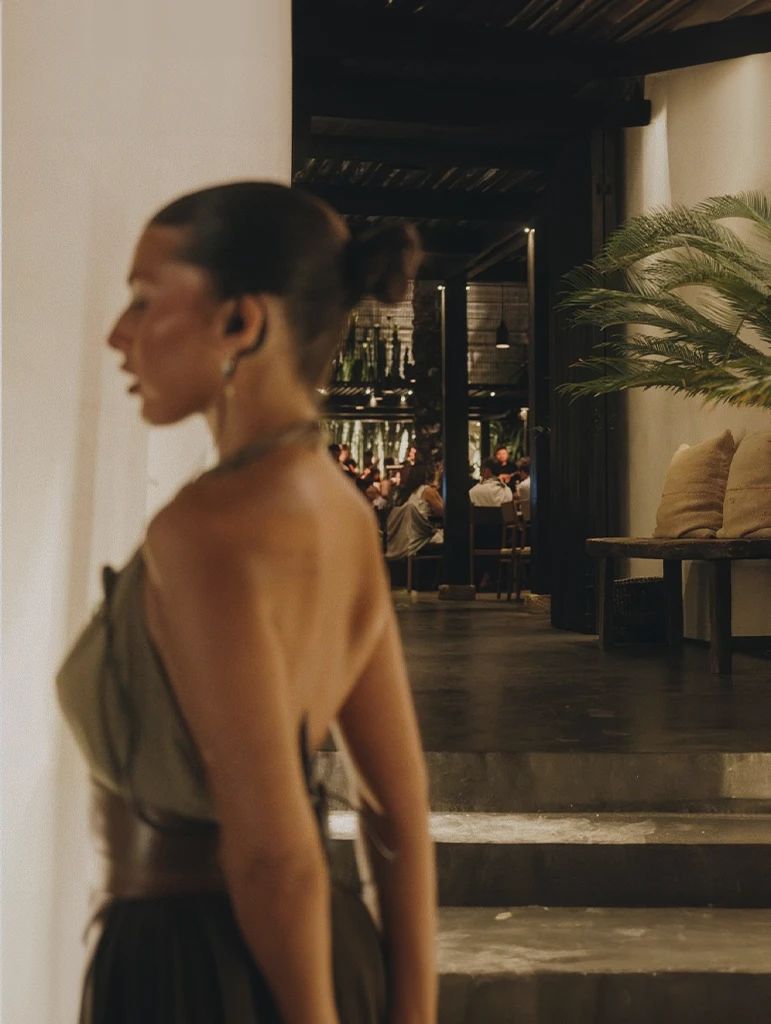 A guest in a silk dress in the candlelit lounge, with diners and palms in the courtyard beyond