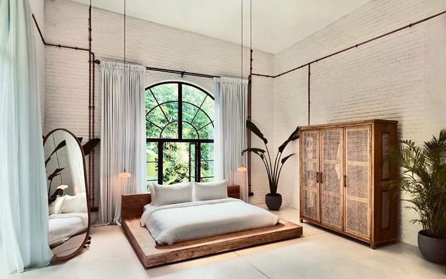 A double-height loft with an arched Crittall window, white-painted brick, exposed copper pipework, a low timber bed and a rattan wardrobe